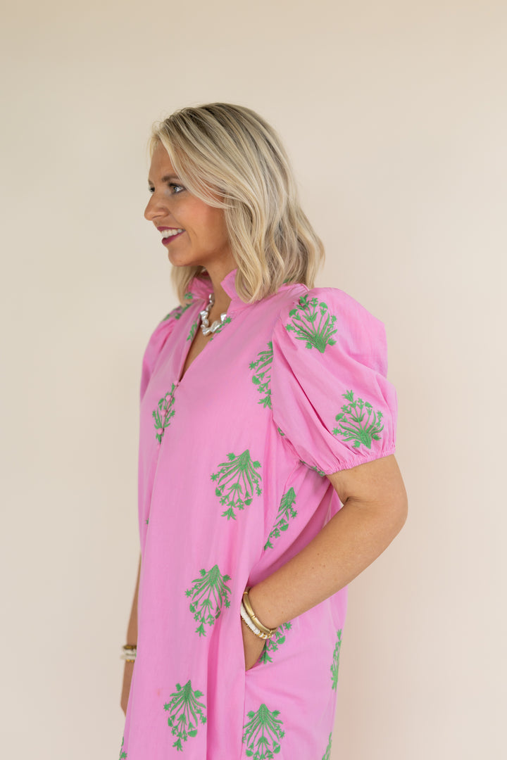 A woman wearing a pink short-sleeve dress with green floral prints, standing against a plain beige background.