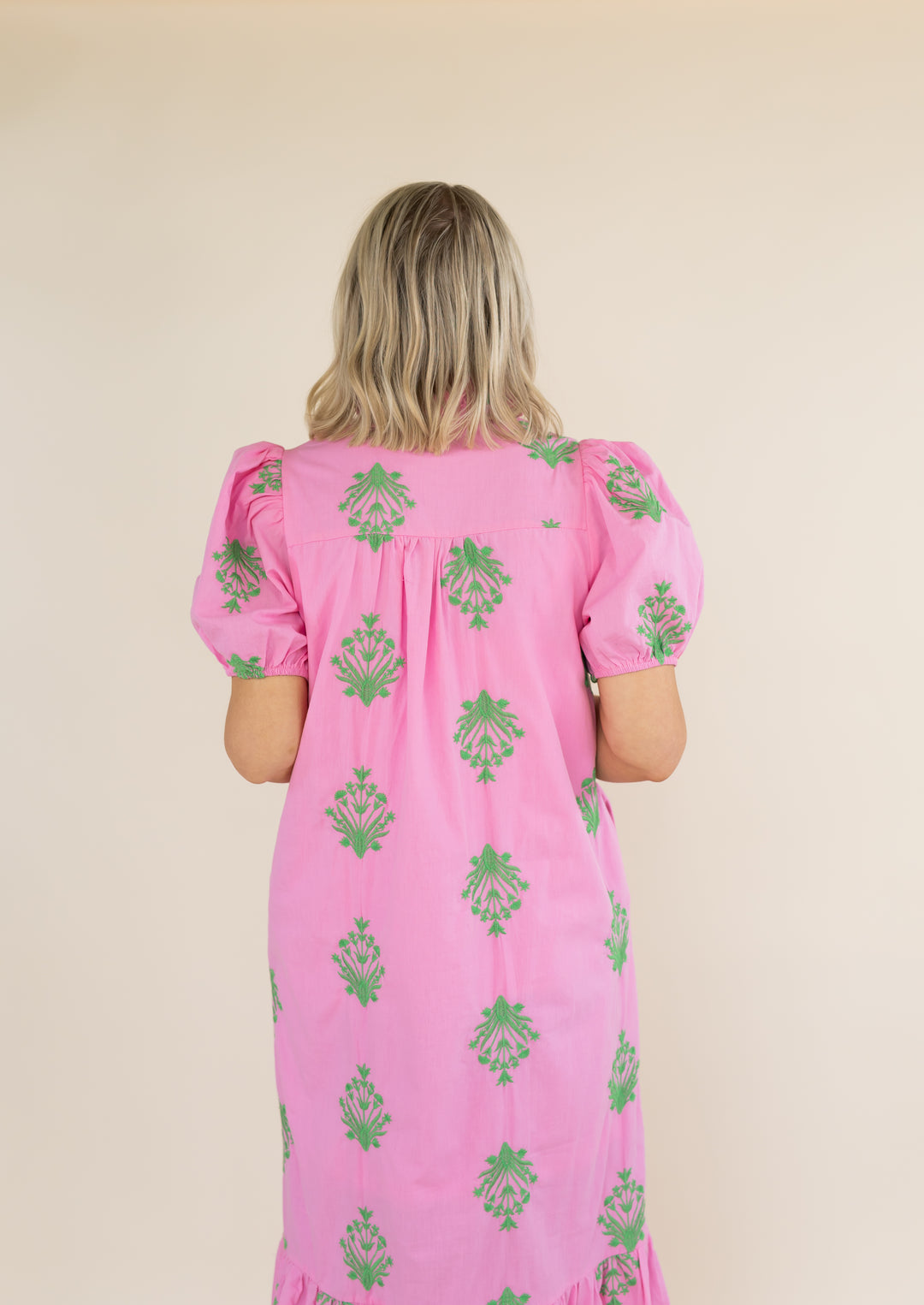 A woman viewed from behind wearing a pink dress with green floral pattern, set against a plain beige background.