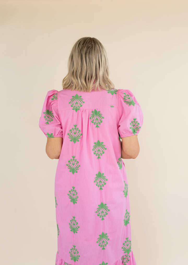 A woman viewed from behind wearing a pink dress with green floral pattern, set against a plain beige background.