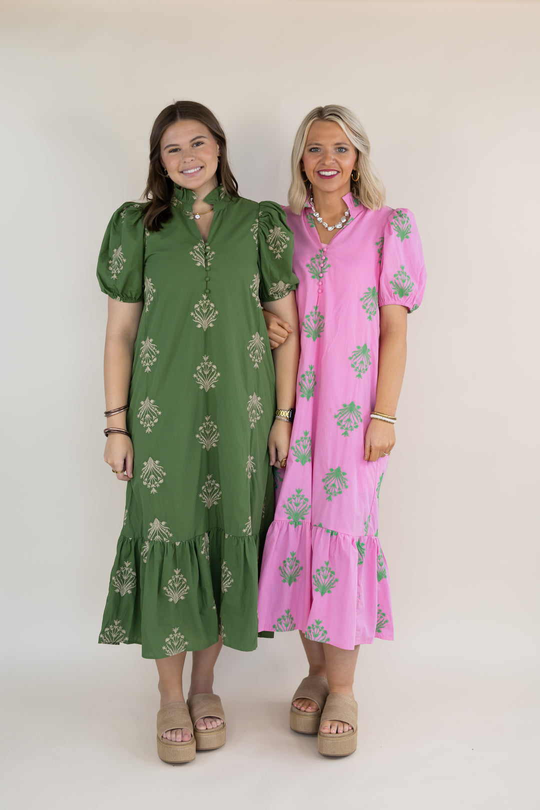 Two women modeling midi-length tiered dresses with puff sleeves and a floral print. The woman on the left wears an olive green dress with beige floral embroidery, and the woman on the right wears a pink dress with olive green floral prints.