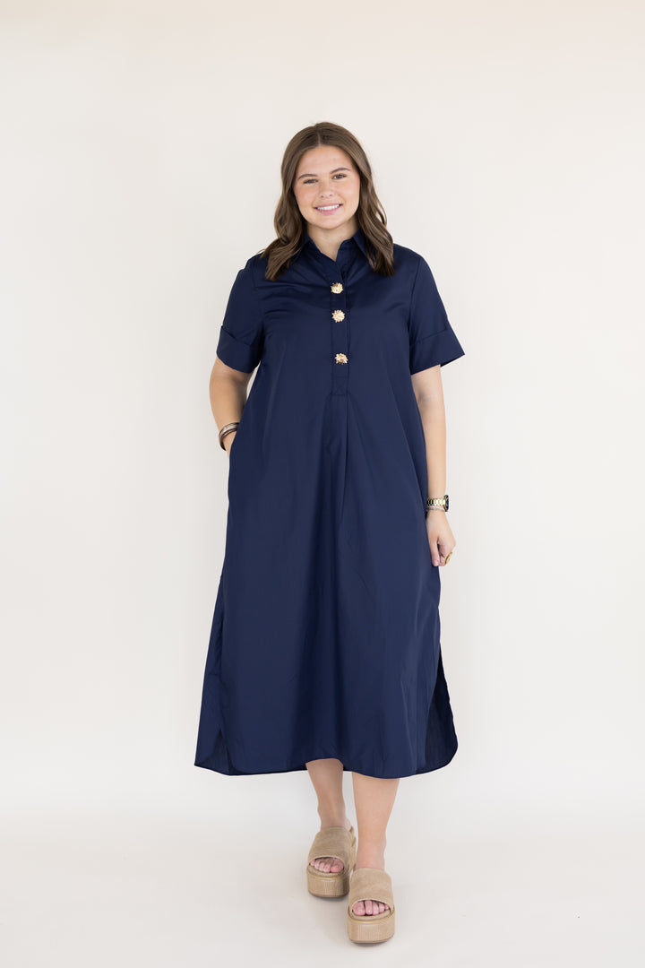 A woman modeling a navy blue midi dress with short sleeves, a button-up front, and gold buttons, standing against a plain white studio background.