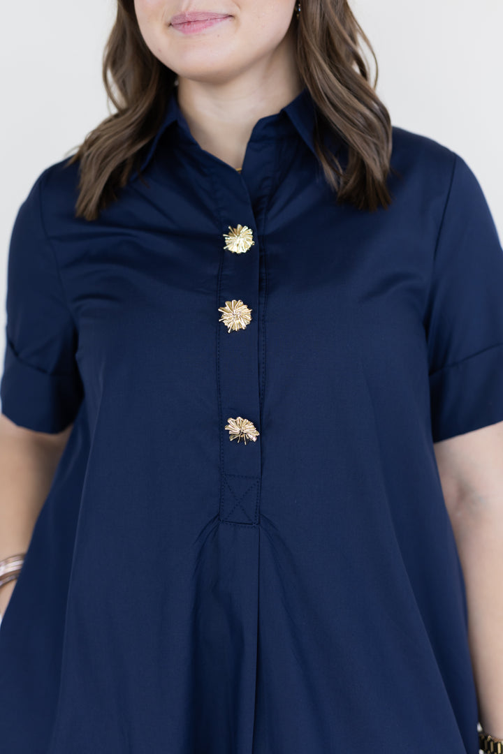 A woman wearing a navy blue short-sleeve collared dress with decorative gold flower-shaped buttons down the front, shown against a plain light background.