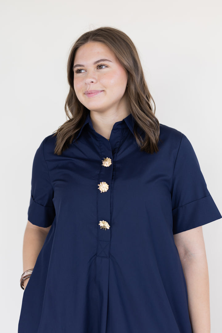 A woman wearing a navy blue short-sleeve button-up dress with decorative gold flower-shaped buttons, photographed against a plain light background.