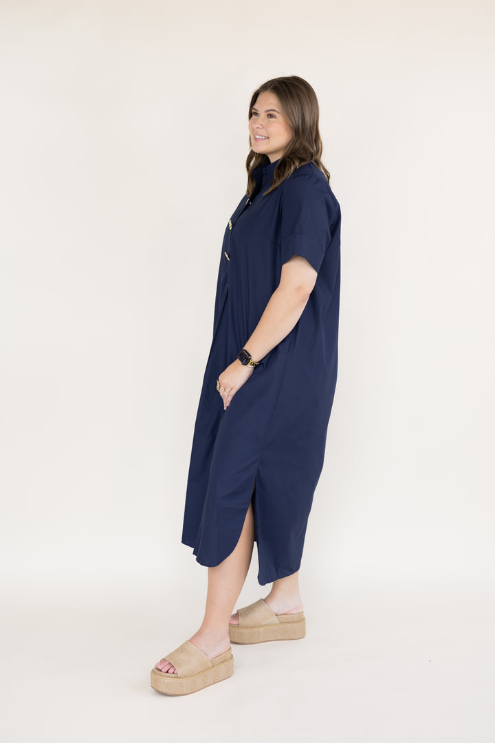 A woman wearing a navy blue short-sleeve midi dress with a high collar and side slits, paired with beige platform sandals, standing against a plain off-white studio background.