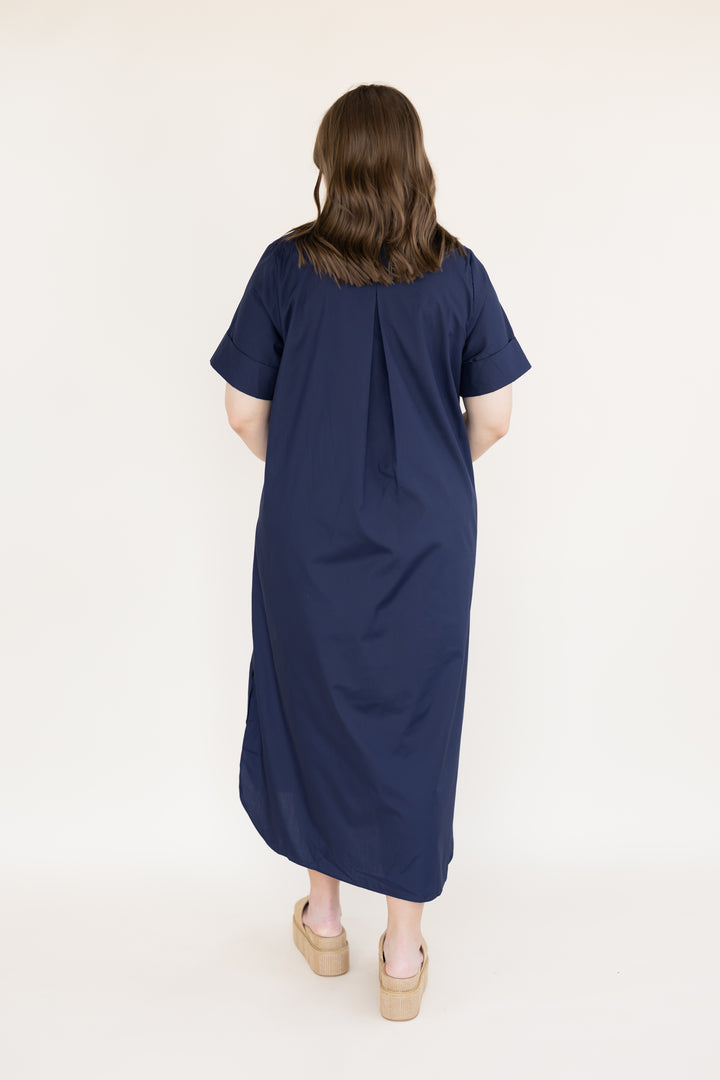 A woman viewed from behind wearing a navy blue short-sleeve midi dress with a gathered back neckline, paired with beige platform sandals, against a plain off-white studio background.
