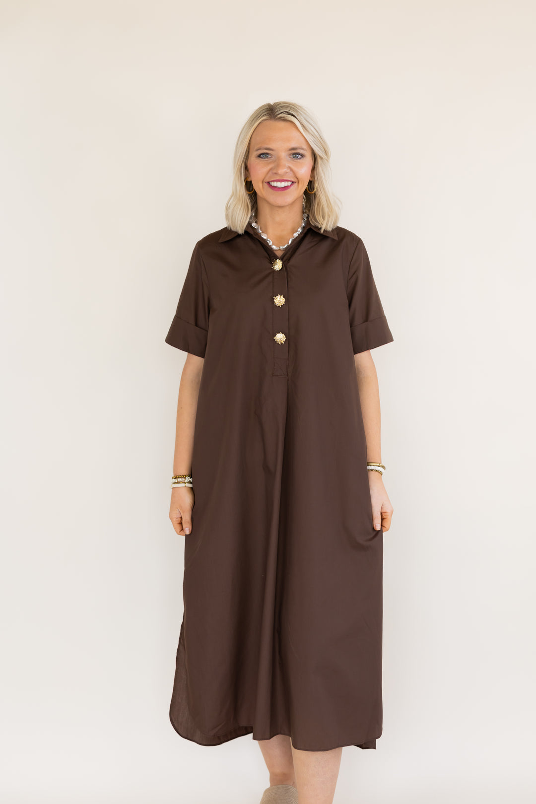 A woman modeling a short-sleeve brown shift dress with gold buttons down the front, standing against a plain off-white background.