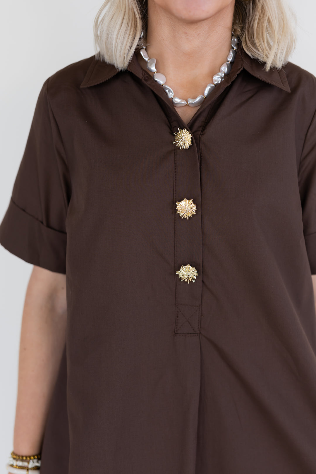 A close-up of a model wearing a dark brown short-sleeve button-up dress with decorative gold flower-shaped buttons, styled with a chunky pearl necklace.