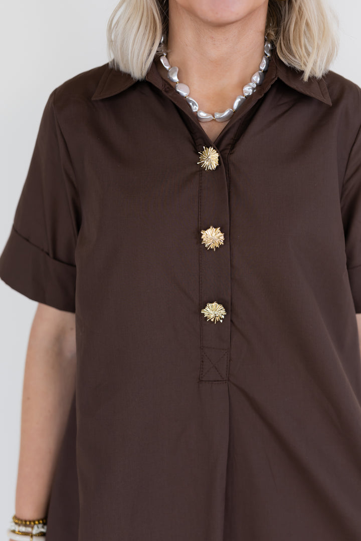 A close-up of a model wearing a dark brown short-sleeve button-up dress with decorative gold flower-shaped buttons, styled with a chunky pearl necklace.