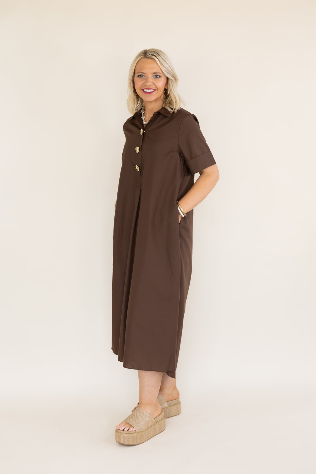 A woman modeling a brown short-sleeve midi dress with gold buttons down the front, standing against a plain off-white studio background.