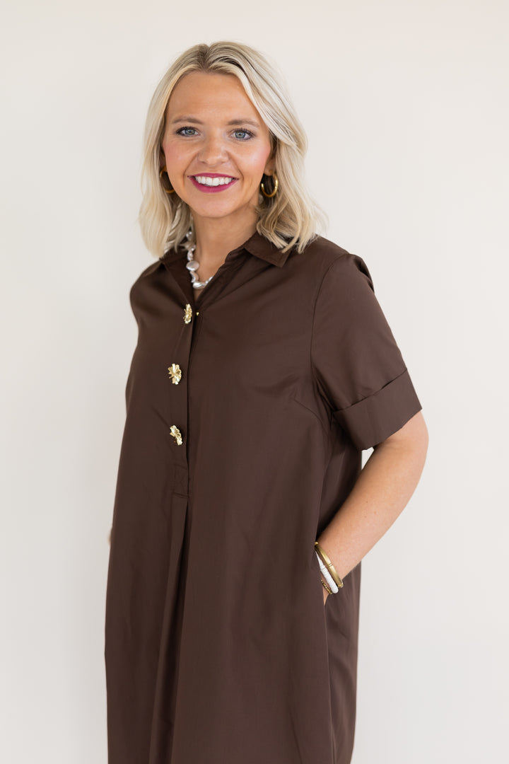 A smiling blonde woman wearing a brown short-sleeve button-front shirt dress with gold-tone buttons, photographed against a plain off-white studio background.