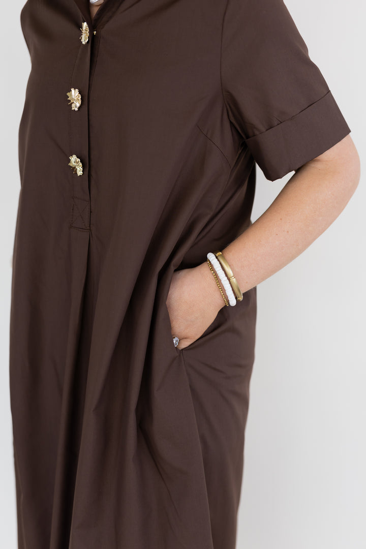 A close-up of a person wearing a dark brown short-sleeve dress with gold decorative buttons and a side pocket, shown against a light gray background.