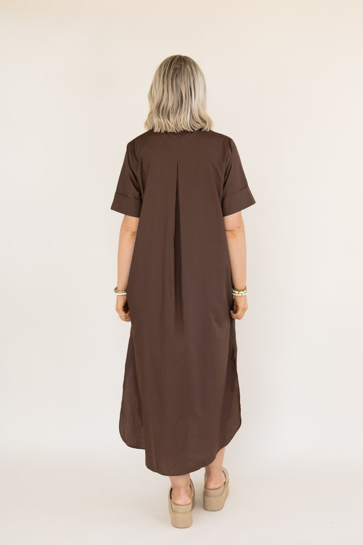 A woman viewed from behind wearing a brown short-sleeve midi dress with a gathered back seam, paired with beige platform sandals, against a plain light beige studio background.