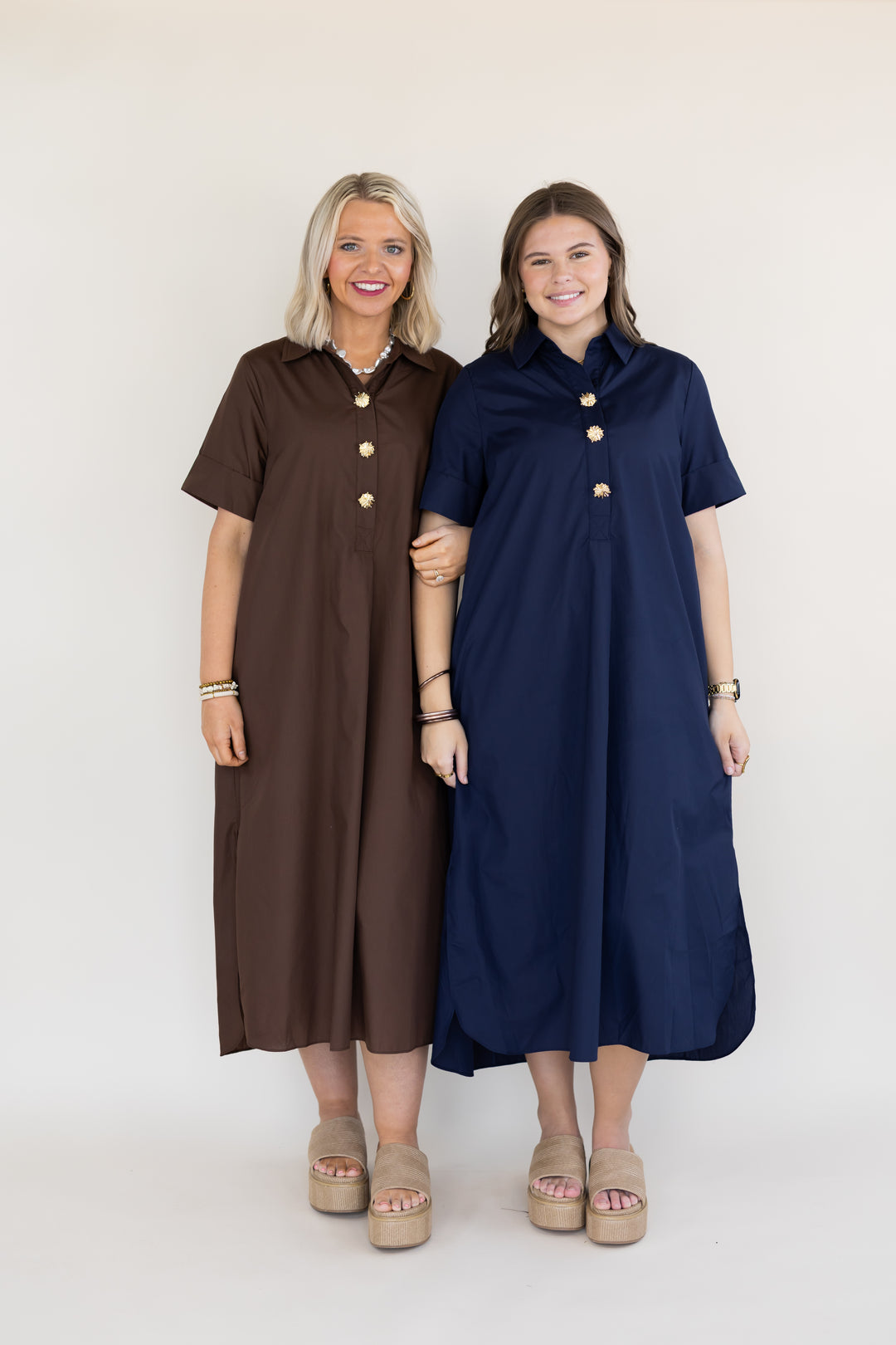 Two women modeling short-sleeve midi dress shirts in brown and navy blue, featuring gold buttons down the front, standing against a plain off-white studio background.