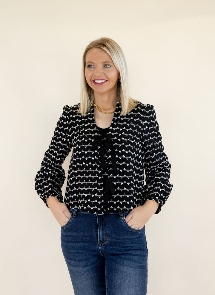 Woman modeling black and white patterned cropped sweater jacket with round neckline, black bow tie closures, long sleeves, and blue jeans