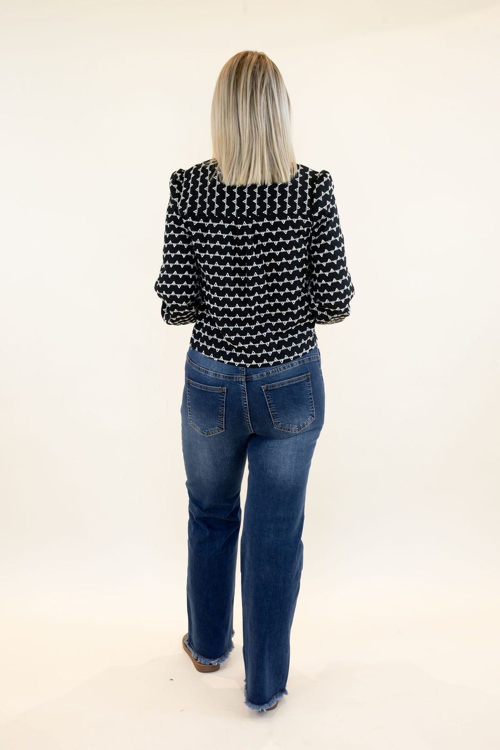 Full-length back view of woman wearing black and white patterned cropped sweater jacket with blue wide leg jeans and black sandals