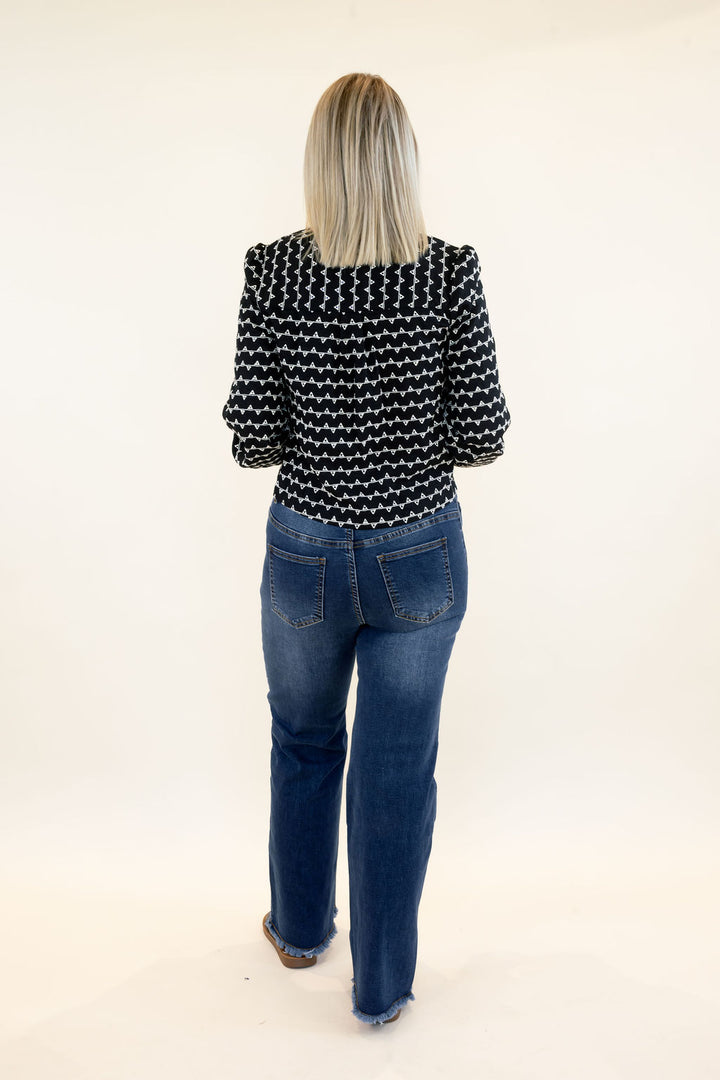 Full-length back view of woman wearing black and white patterned cropped sweater jacket with blue wide leg jeans and black sandals
