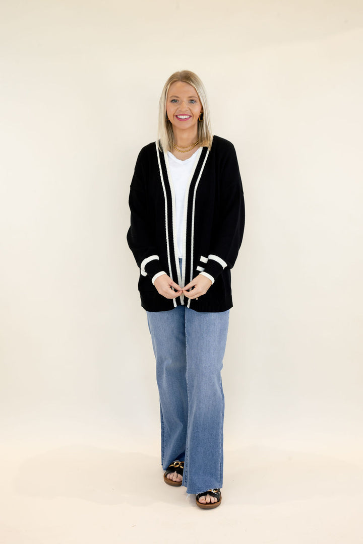Woman modeling black open-front cardigan with contrasting white trim, long sleeves, front patch pockets, and relaxed fit styled with a white top and light wash wide leg jeans