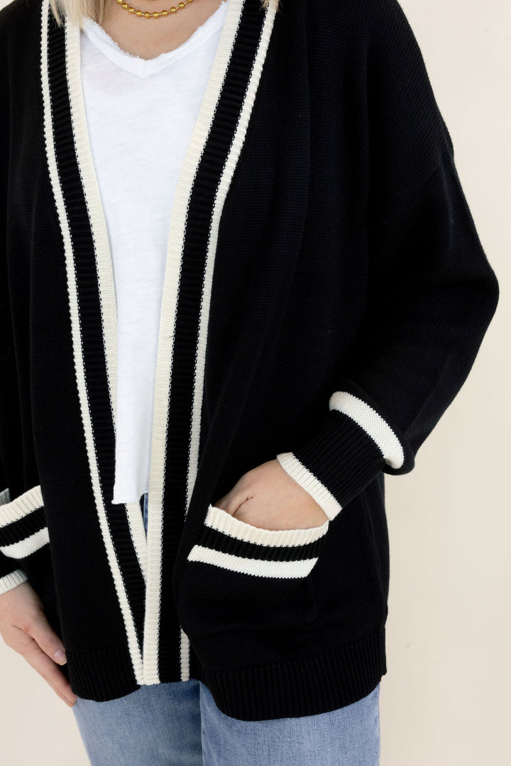 Close-up of black cardigan highlighting the white contrast trim, open-front design, long sleeves with striped cuffs, and front patch pockets