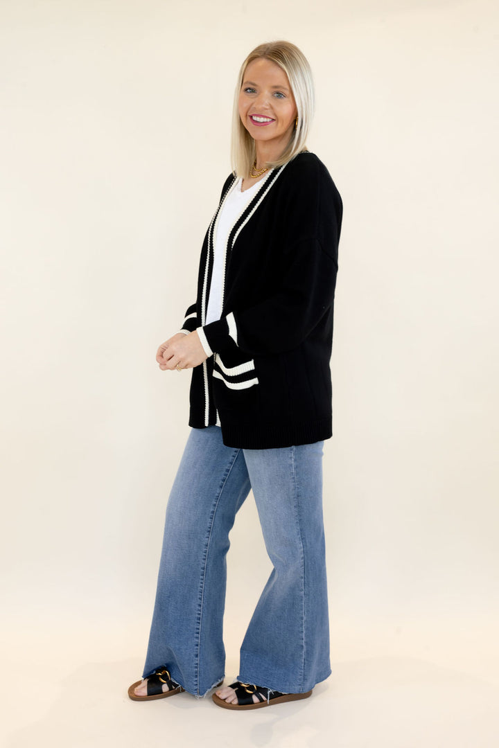Side view of woman wearing black cardigan with white contrast trim, open-front design, and long sleeves styled with blue wide leg jeans and black sandals
