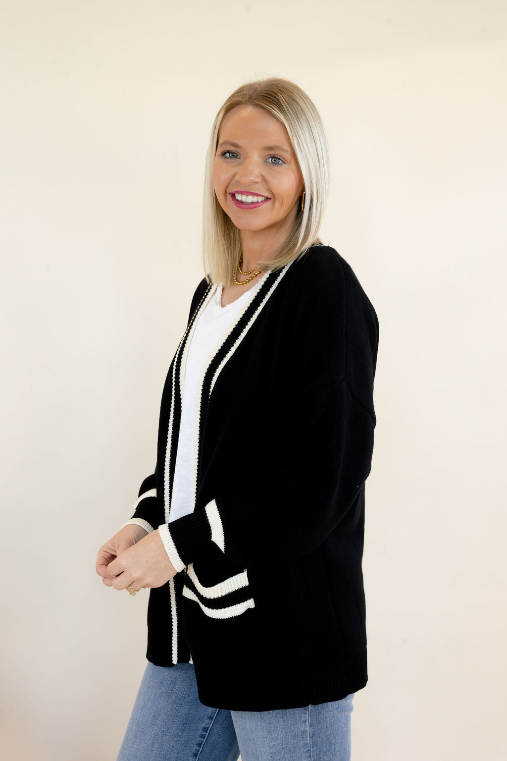 Woman modeling black open-front cardigan with white trim, contrasting cuff details, and front patch pockets styled with a white V-neck top and blue jeans