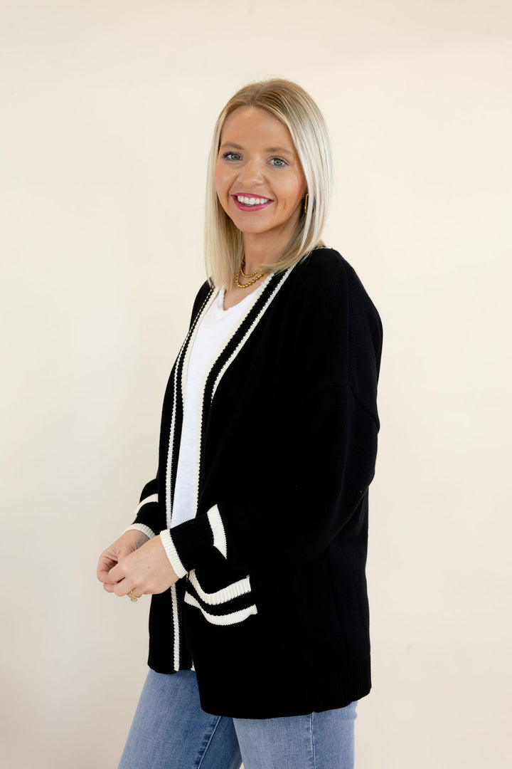 Woman modeling black open-front cardigan with white trim, contrasting cuff details, and front patch pockets styled with a white V-neck top and blue jeans