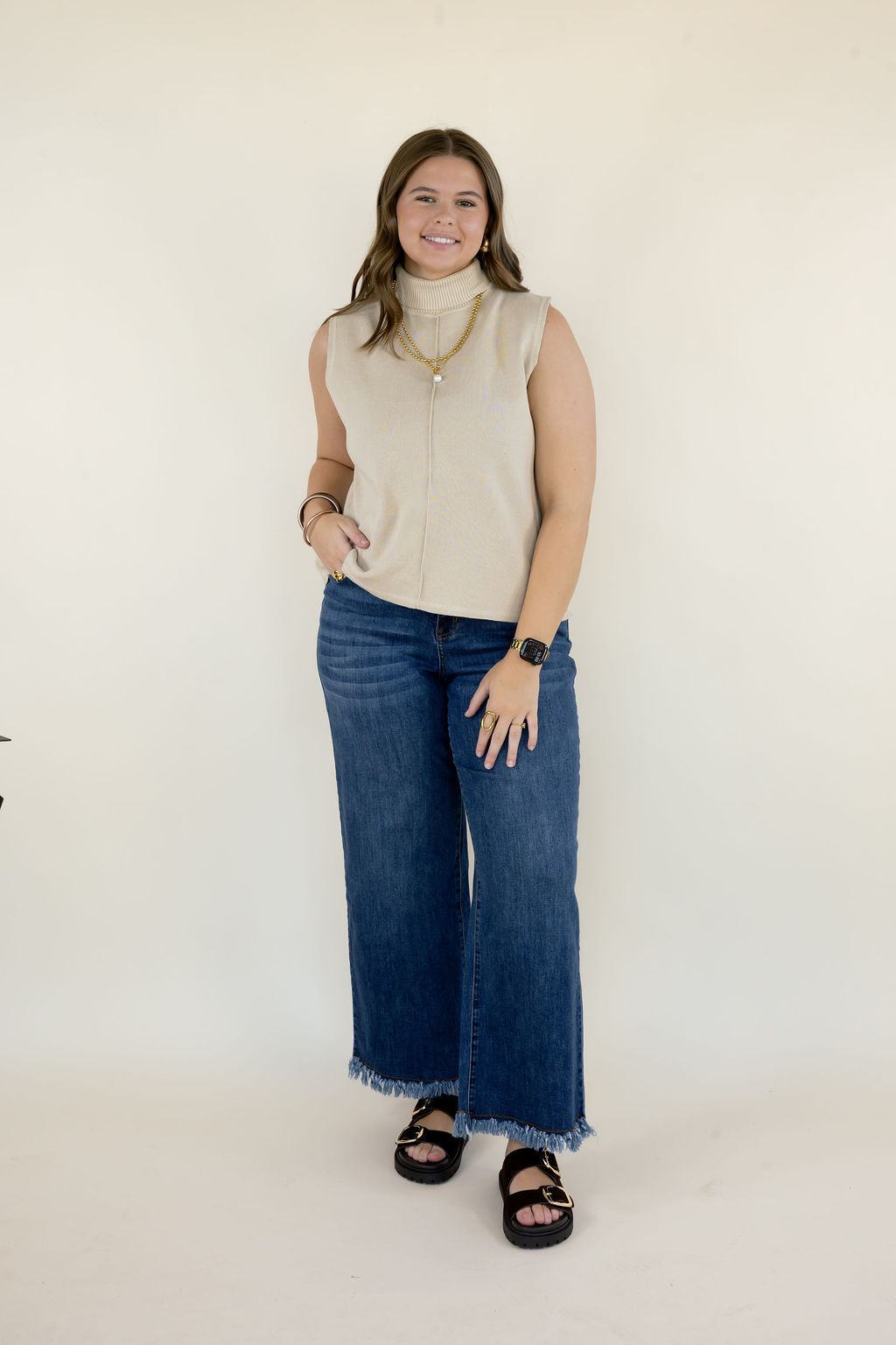 Woman modeling cream sleeveless mock neck top with high neckline, relaxed silhouette, blue wide leg jeans, and black sandals