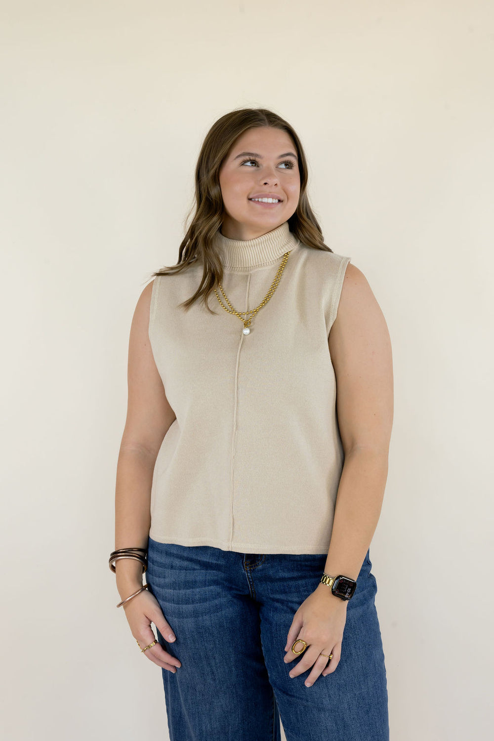 Woman modeling cream sleeveless mock neck sweater top featuring a high neckline, relaxed fit, and gold layered necklaces styled with blue jeans