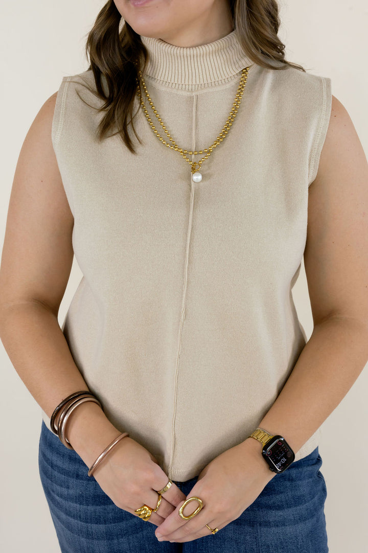 Close-up of cream sleeveless mock neck knit top showing the center front seam, smooth knit texture, and gold layered necklaces