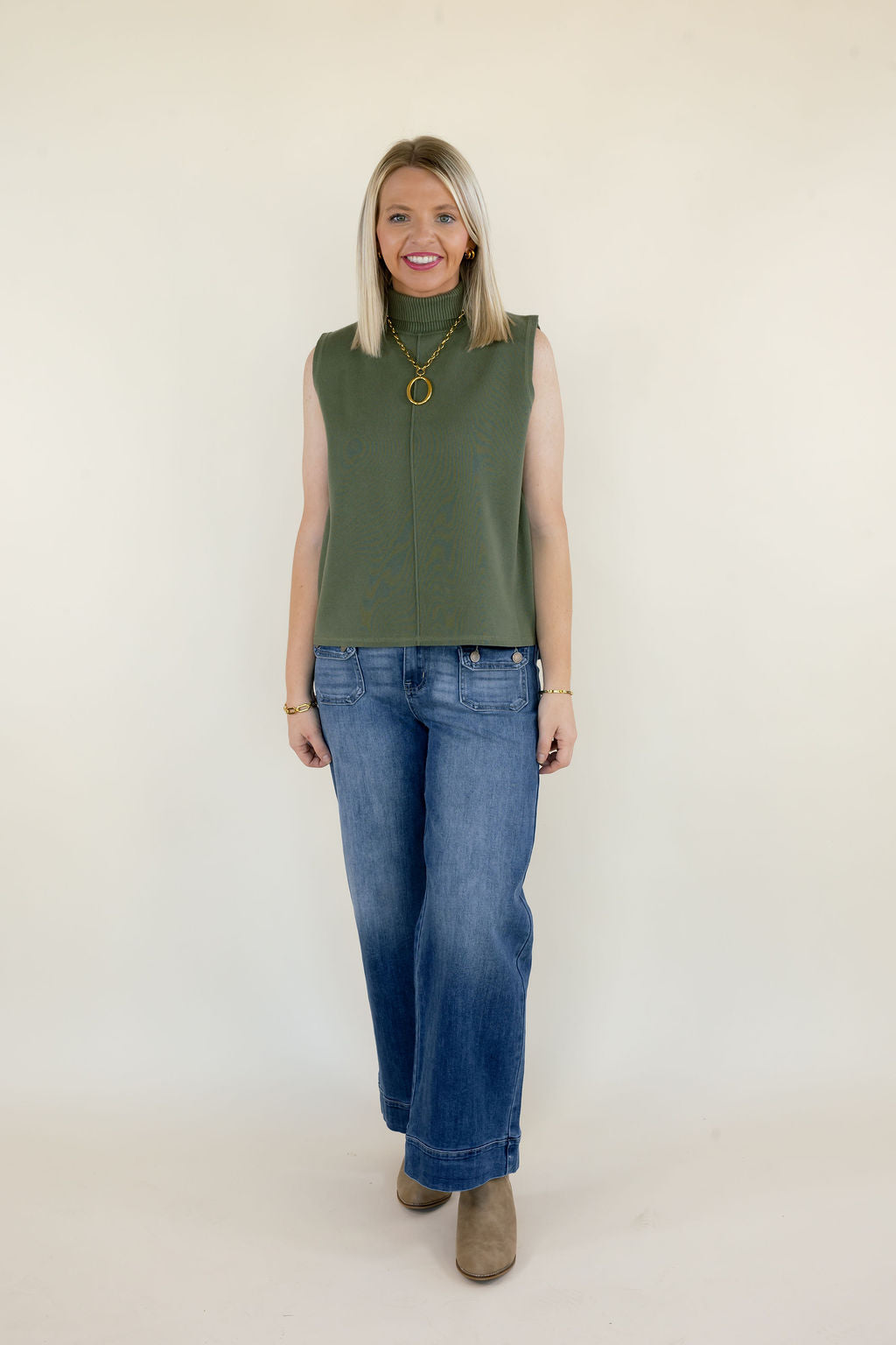 Full-length view of woman wearing olive green sleeveless mock neck knit top with relaxed fit, blue wide leg jeans, and neutral ankle boots
