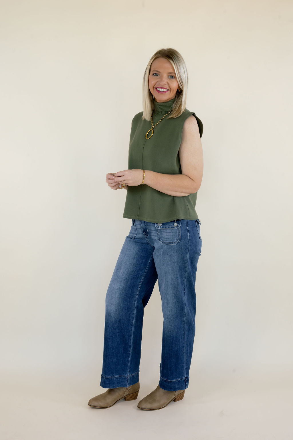 Side view of woman modeling olive green sleeveless mock neck top with relaxed fit, high neckline, and blue wide leg jeans