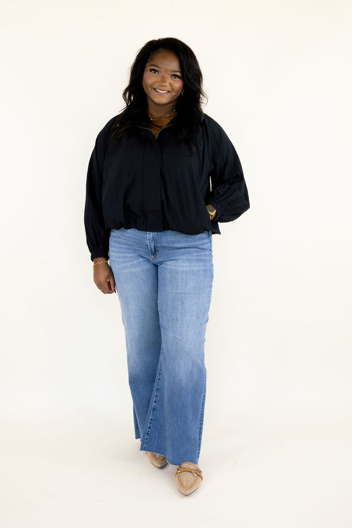 Woman wearing black collared balloon sleeve blouse with relaxed silhouette, gathered detailing, and light wash wide leg jeans