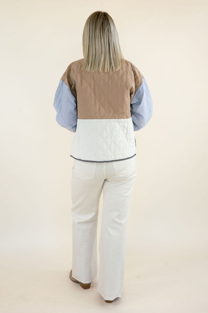 Back view of woman wearing camel brown, ivory, and light blue colorblock quilted jacket showing the relaxed silhouette and white wide leg pants