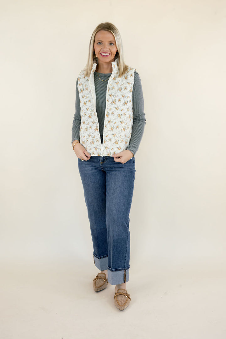 Woman wearing ivory floral print quilted vest with sage green long sleeve top, cuffed straight leg jeans, and tan flats
