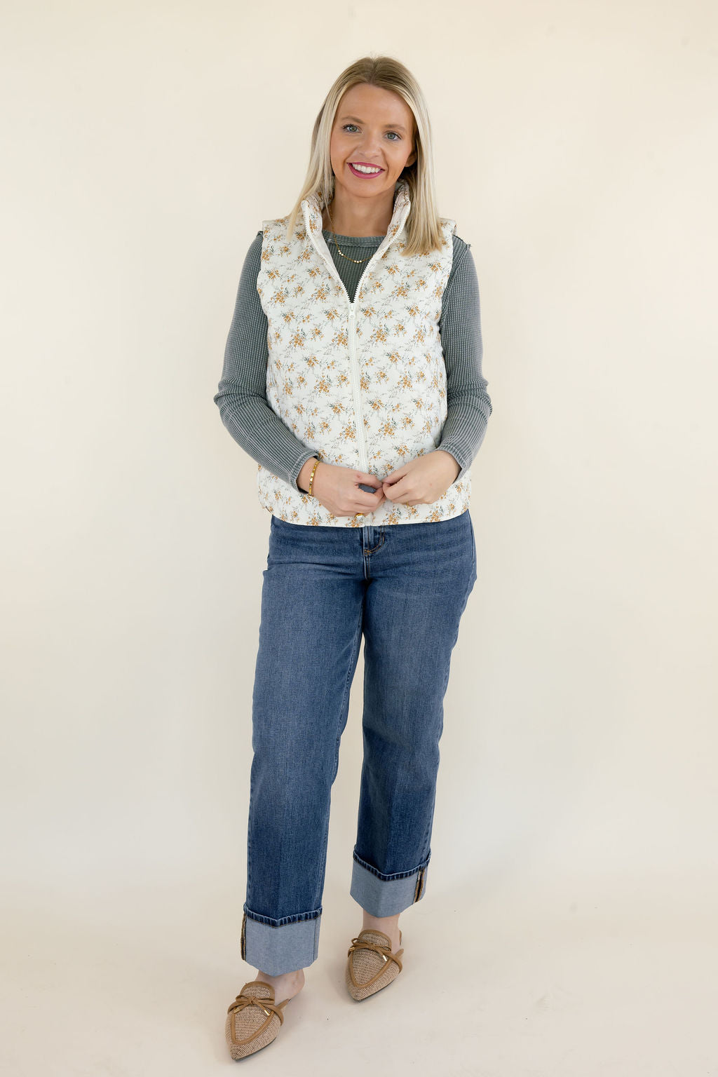 Woman modeling ivory floral quilted vest with small golden botanical print, high collar, and zip front layered over a sage green ribbed top with cuffed blue jeans