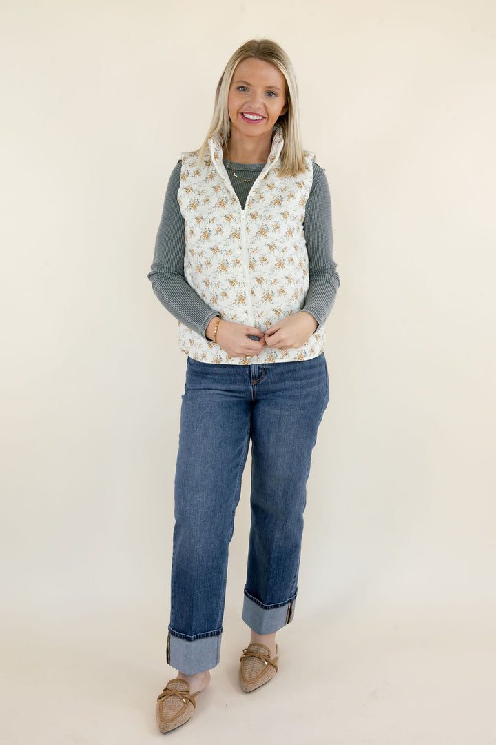 Woman modeling ivory floral quilted vest with small golden botanical print, high collar, and zip front layered over a sage green ribbed top with cuffed blue jeans