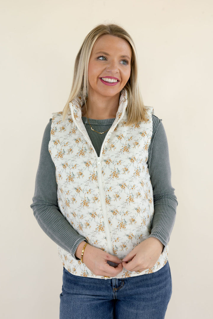 Front view of ivory floral quilted puffer vest featuring delicate golden floral print, stand collar, sleeveless design, and zip-up closure