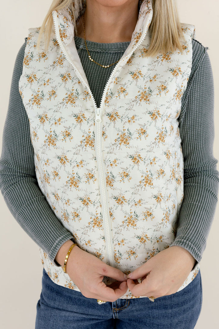 Close-up of ivory floral print quilted vest showing the zip-up front, delicate golden flowers, high collar, and lightly padded construction