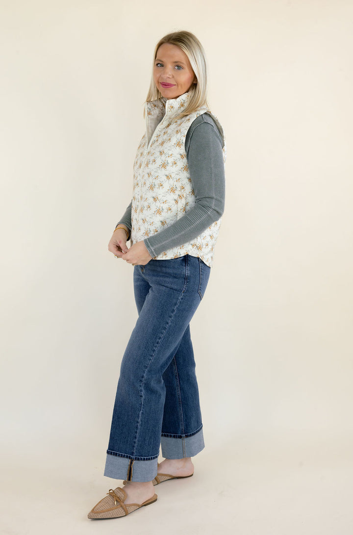 Side view of woman wearing ivory floral quilted vest with sage green ribbed long sleeve top, cuffed blue jeans, and tan flats