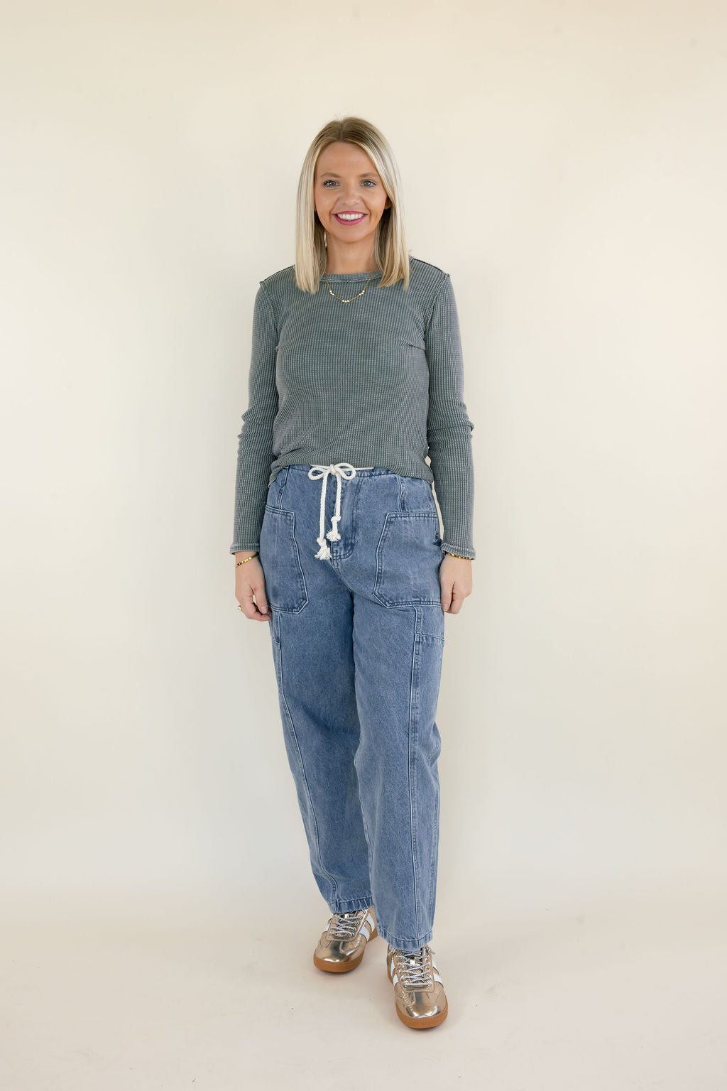 Woman modeling light wash denim cargo pants with white drawstring waistband, oversized utility pockets, and relaxed straight-leg silhouette styled with a gray ribbed sweater and metallic sneakers