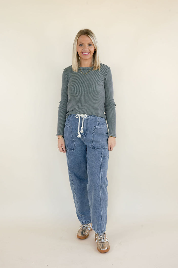 Woman modeling light wash denim cargo pants with white drawstring waistband, oversized utility pockets, and relaxed straight-leg silhouette styled with a gray ribbed sweater and metallic sneakers