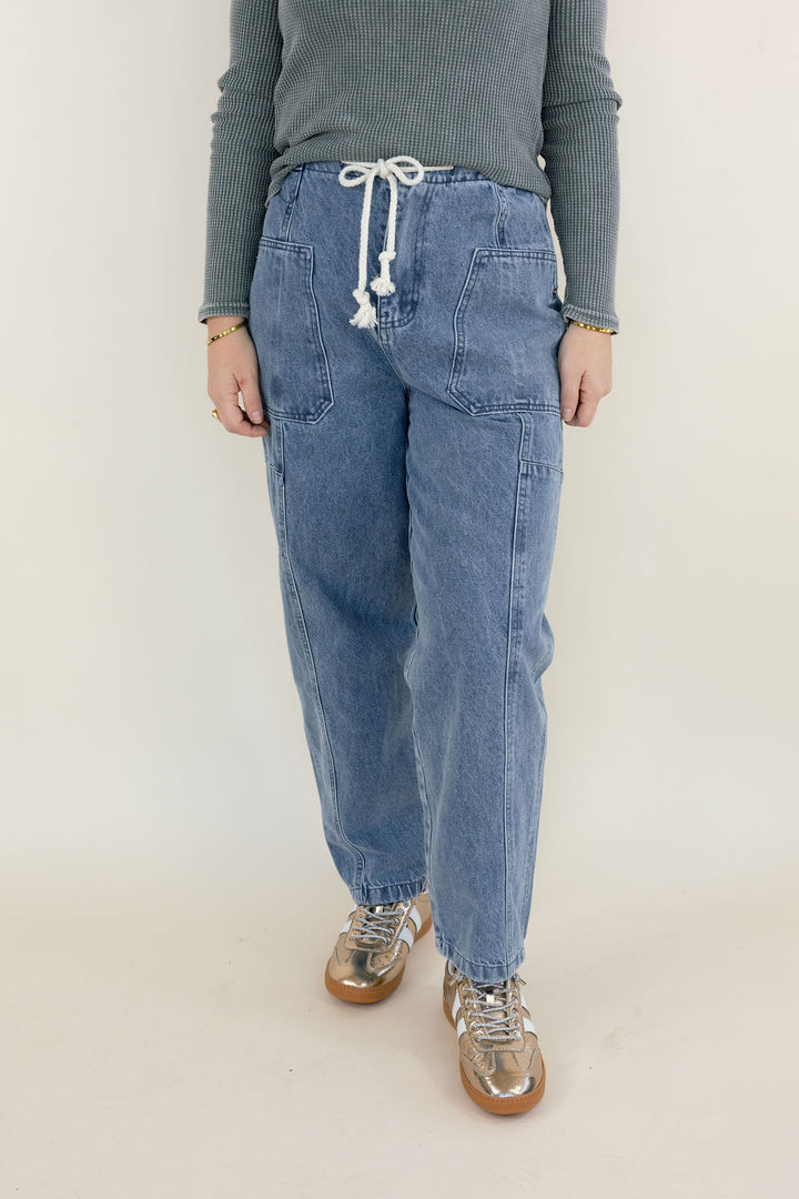 Front view of light wash denim cargo pants showing adjustable white drawstring waist, large front patch pockets, relaxed fit, and full-length legs