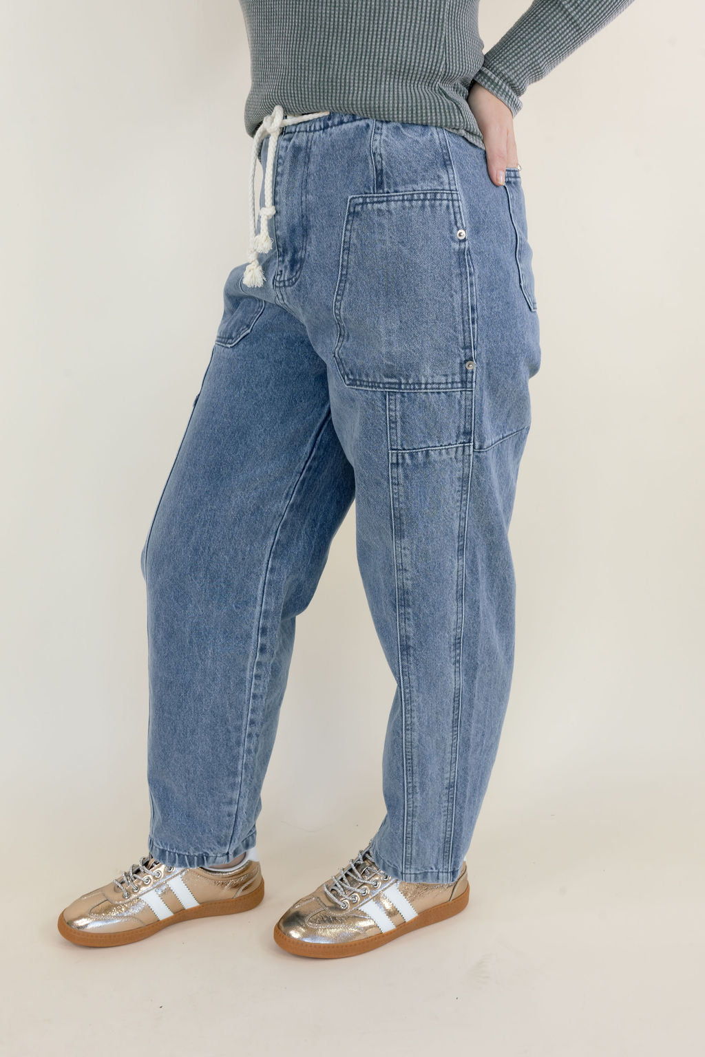 Side view of woman modeling light wash denim cargo pants featuring oversized utility patch pockets, white drawstring waistband, relaxed straight legs, and metallic sneakers