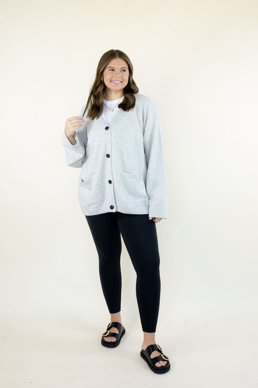 Full-length view of women's light gray knit cardigan sweater with black button-front closure and patch pockets, paired with black leggings and casual sandals.