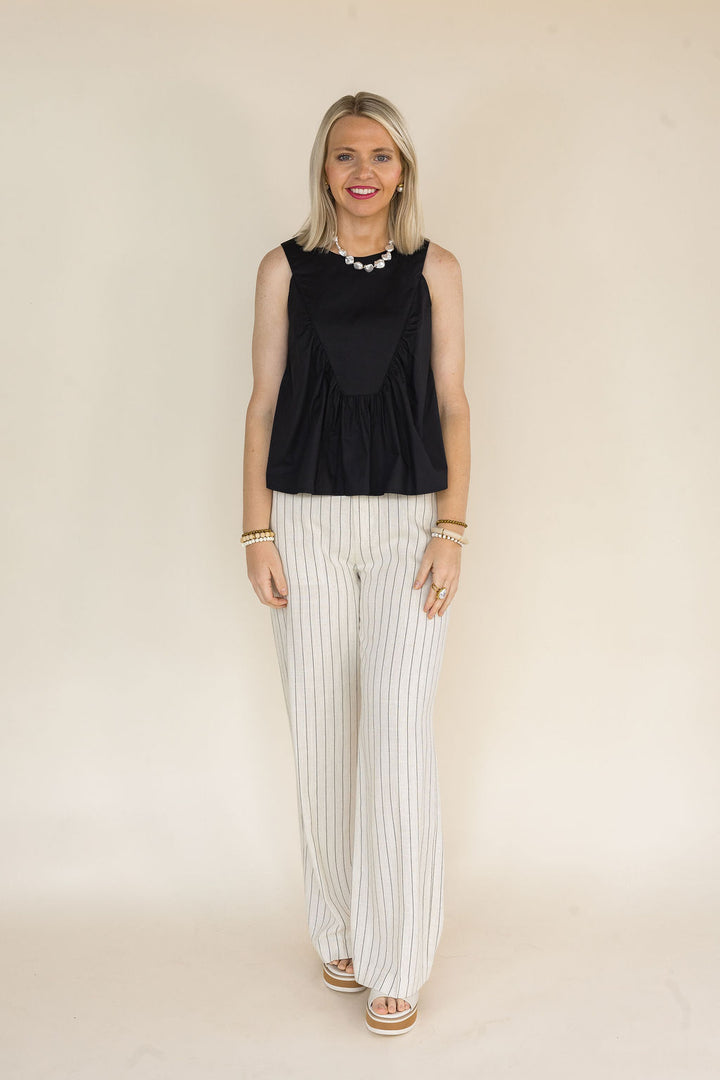 A woman modeling a sleeveless black peplum top paired with cream wide-leg trousers featuring thin vertical black stripes, set against a plain beige studio background.