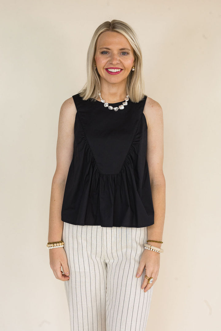 Woman wearing a black sleeveless ruffle blouse paired with cream pinstripe wide-leg pants, pearl jewelry, and stacked bracelets for a polished casual outfit.
