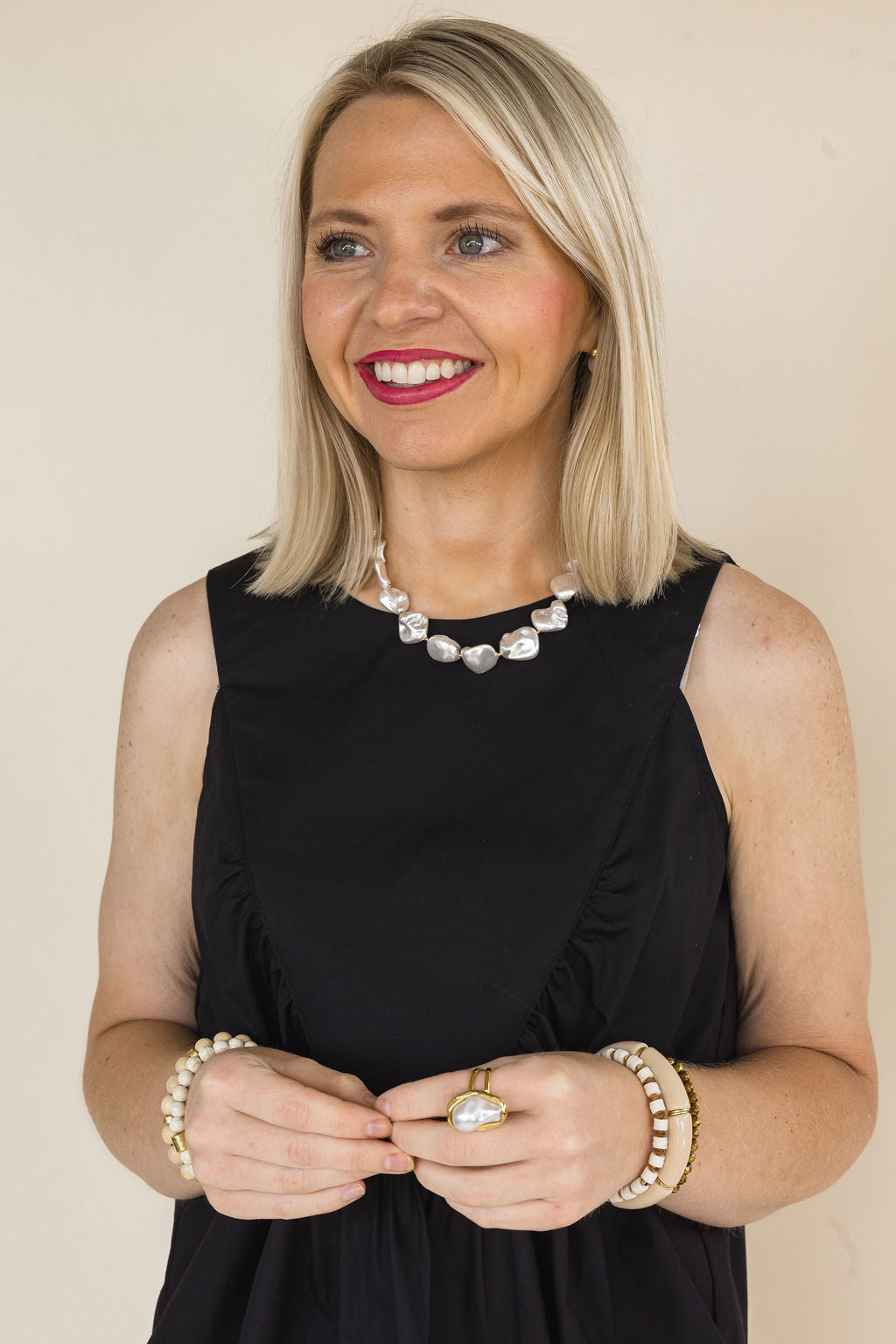 Woman wearing a black sleeveless ruffle detail blouse with a gathered front yoke, styled with a pearl statement necklace, stacked bracelets, and a gold statement ring.