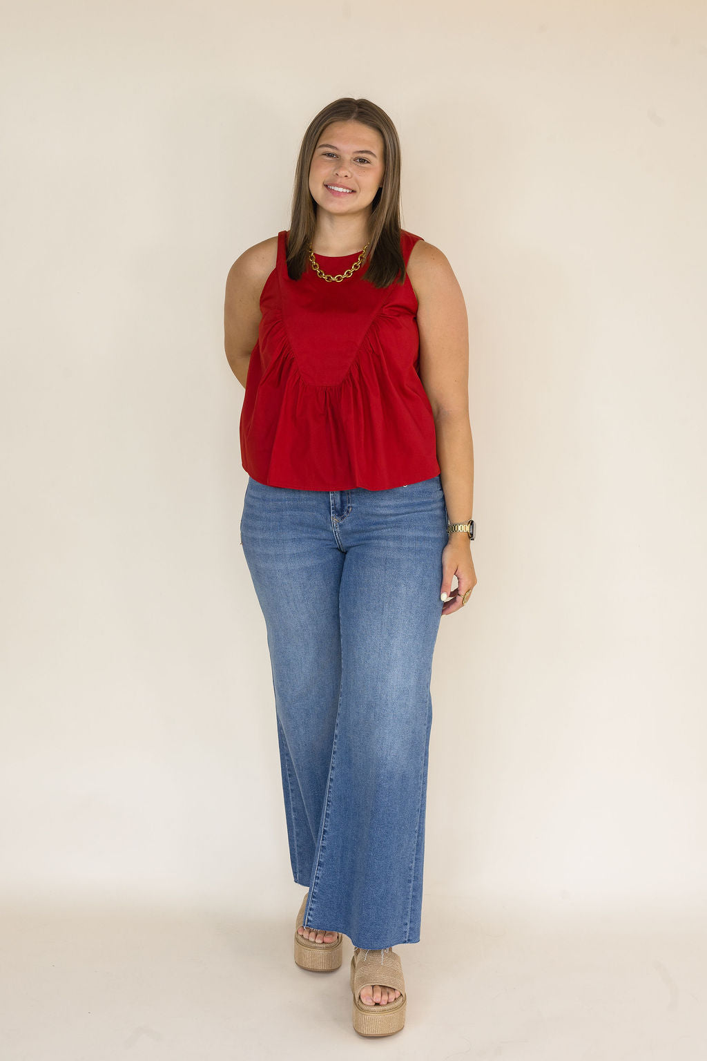 A woman modeling a sleeveless garnet peplum top with blue wide-leg jeans and beige platform sandals, standing against a plain beige studio background.
