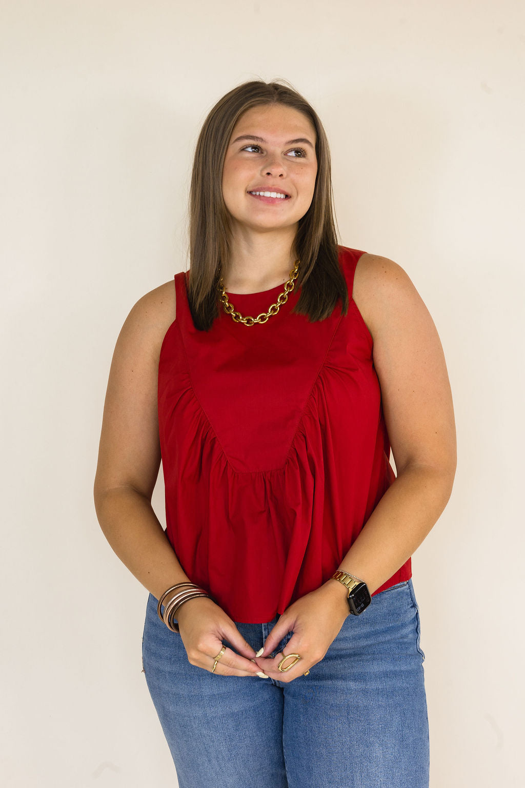 A woman wearing a sleeveless garnet top and blue jeans, standing against a plain light beige background.