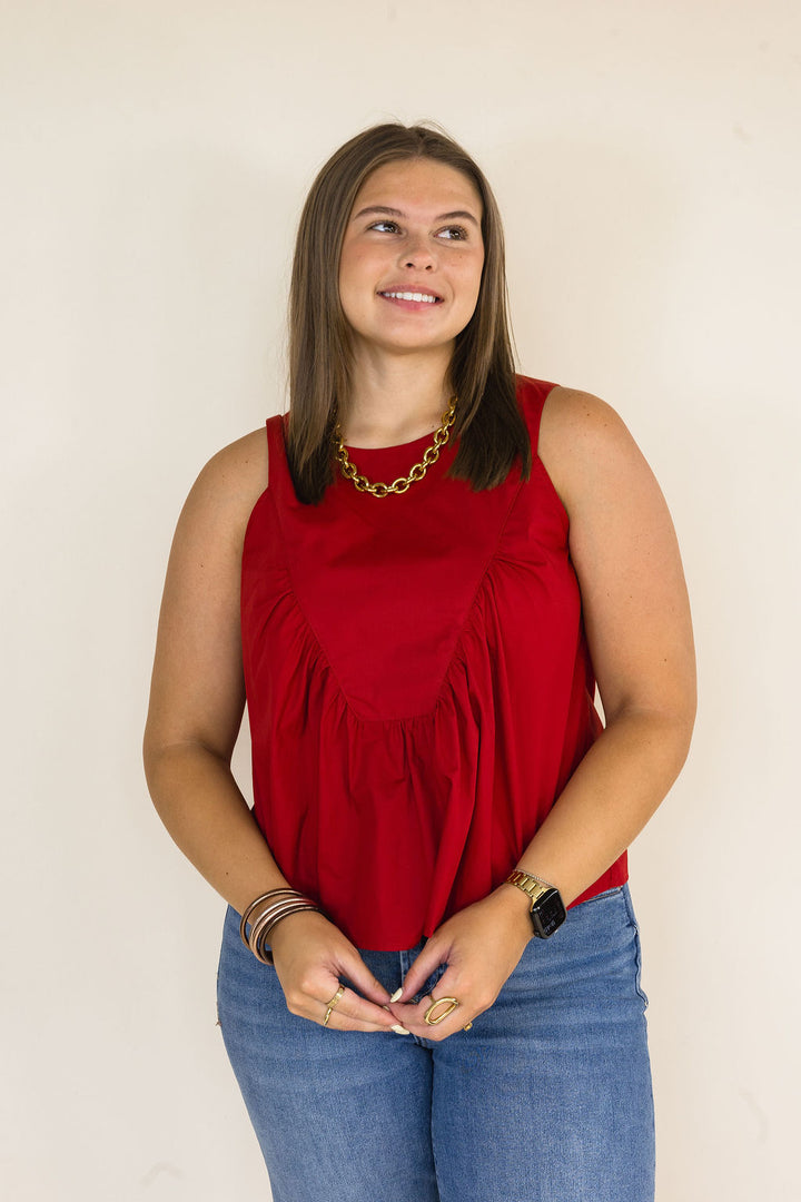 A woman wearing a sleeveless garnet top and blue jeans, standing against a plain light beige background.