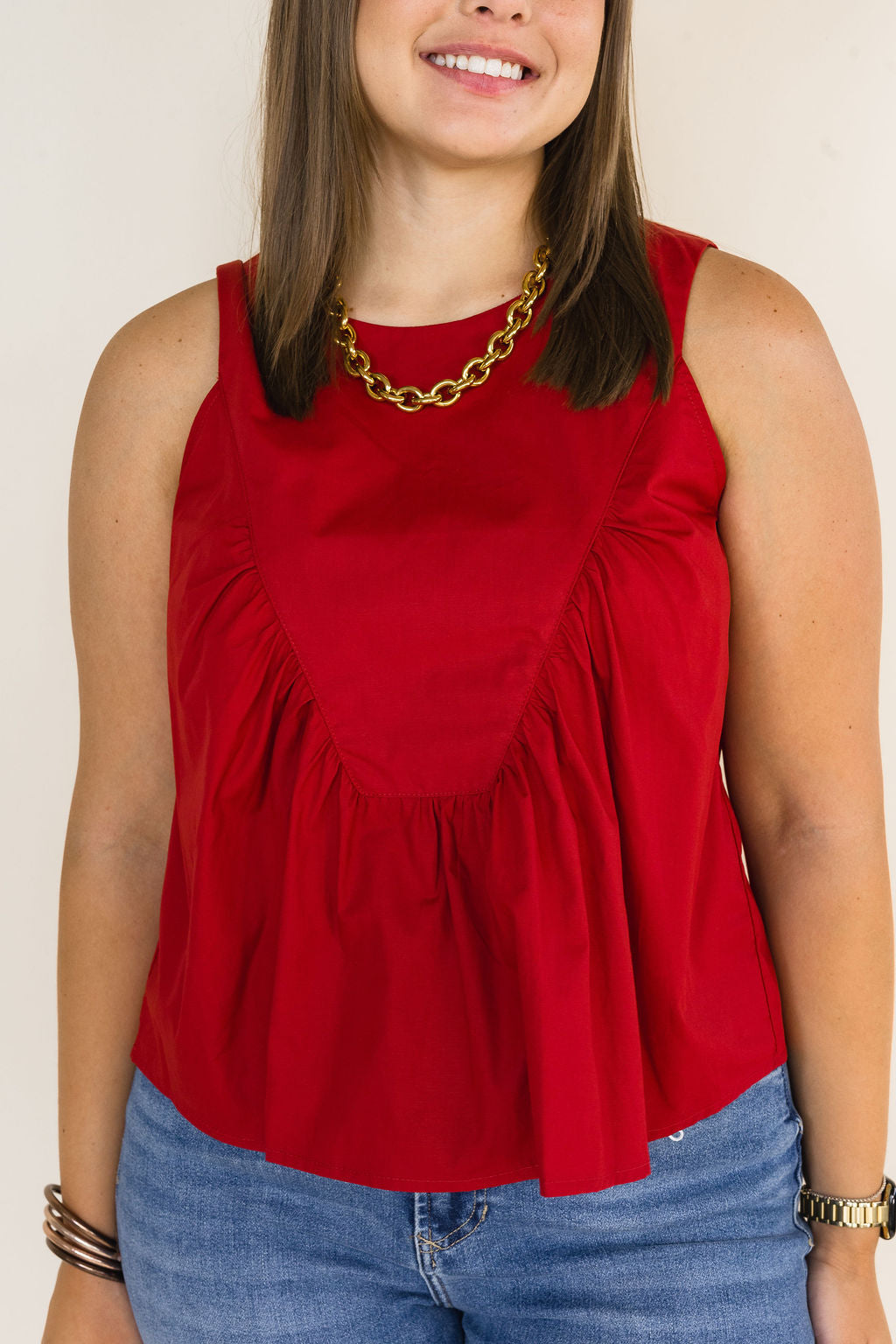 Woman wearing a garnet sleeveless ruffle blouse featuring a gathered front yoke, styled with blue jeans, a chunky gold chain necklace, and gold bracelets.
